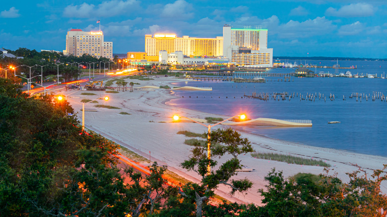 A white sand beach with boats on its waters, and buildings with their lights on in the evening.