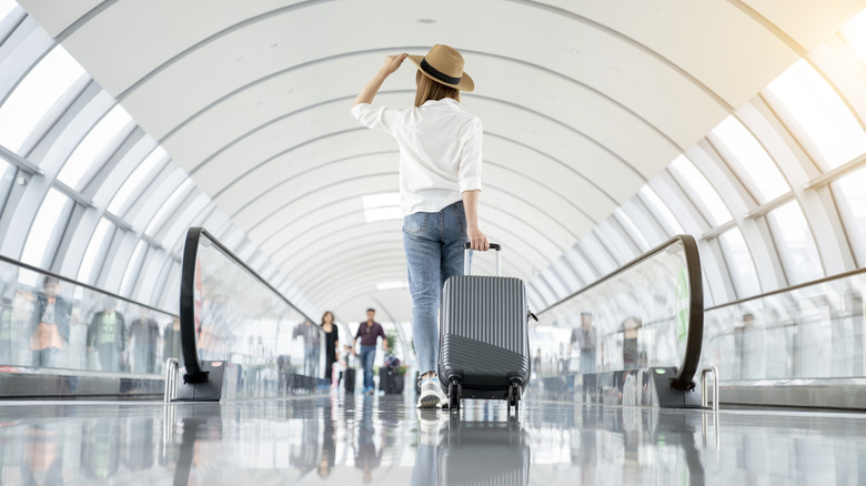 A woman walking with her rolling suitcase behind her in a modern airport terminal.