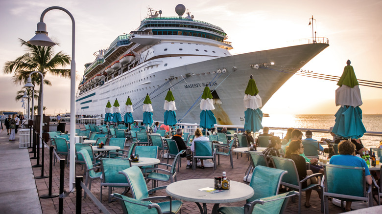 fancy white tables and blue chairs set around the dock of a docked cruise ship with palm trees and sunset in background.
