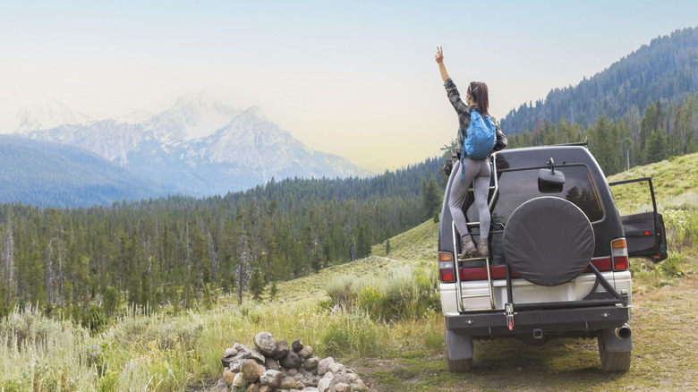 A woman holds up a peace sign while standing on her car, overlooking a mountain range.