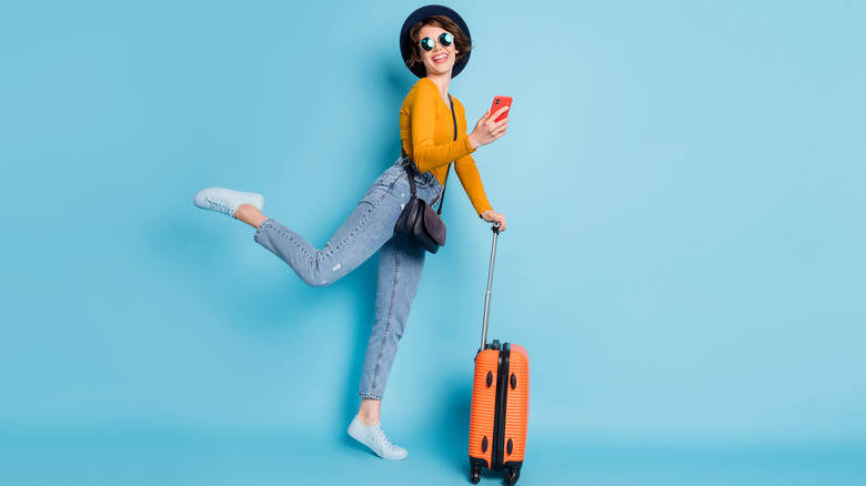 A woman wearing white sneakers with a leg kicked back, holding a phone, leaning on a small luggage