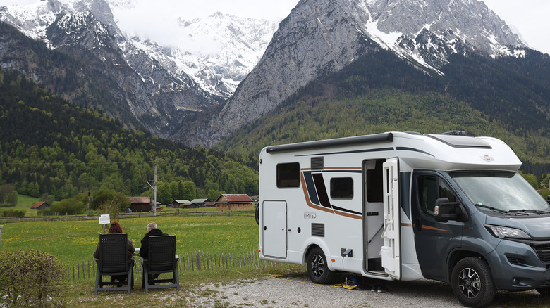 Two people sitting next to parked RV with mountain view in background