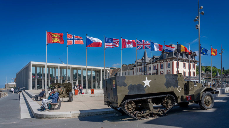 A military tank in front of buildings and the flags of multiple countries.