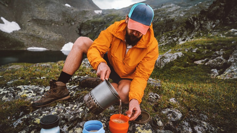 A male hiker pours hot water into an orange cup next to an alpine lake