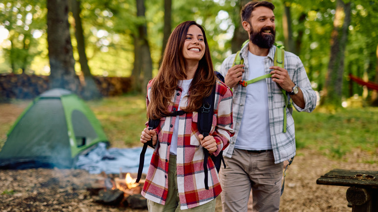 Smiling hikers with backpacks stroll through a lush forest, enjoying their outdoor adventure near a cozy campsite
