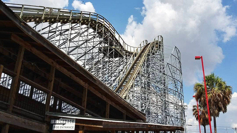 A roller coaster near palm trees with a blue cloudy sky overhead.