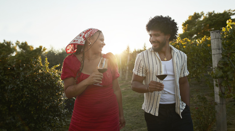 Smiling couple with red wine walking amidst plants in farm during sunset