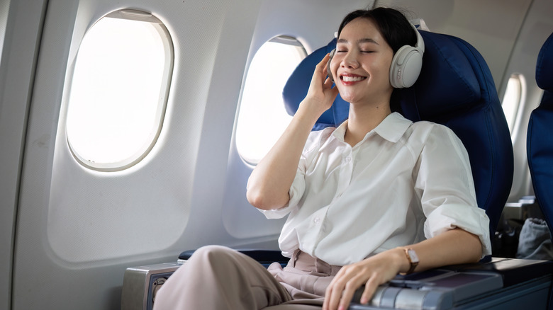 An Asian woman sitting in an airplane seat with headphones on while smiling with her eyes closed