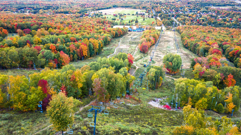 A chair lift going up a small ski hill with fall trees all around and in the distance