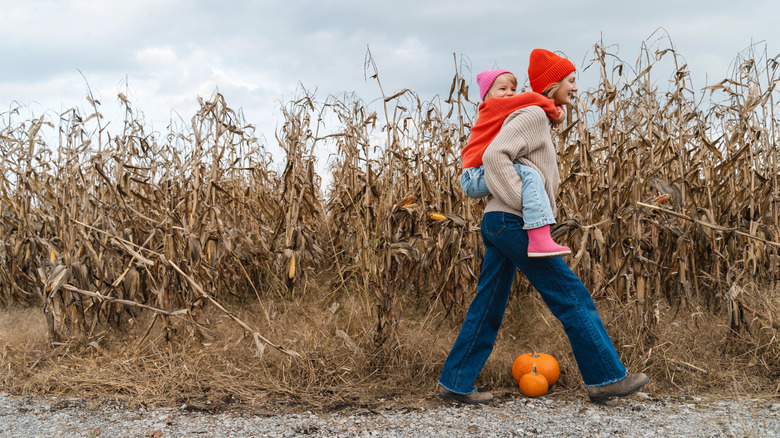 A mother carrying her young daughter by autumn corn stalks