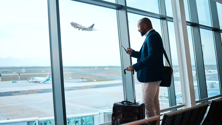 Man holding phone close to airport window and view of plane taking off
