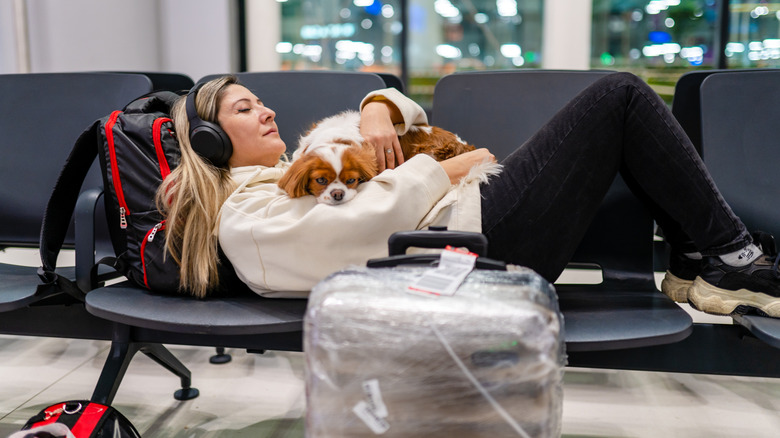 A person lying on airport seats surrounded by bags and cuddling a small dog