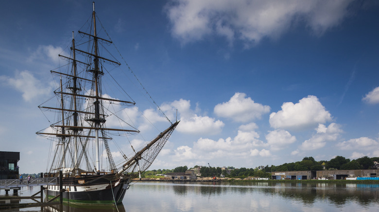 An old sail ship on a river with sun, clouds, and the opposing shoreline in the distance