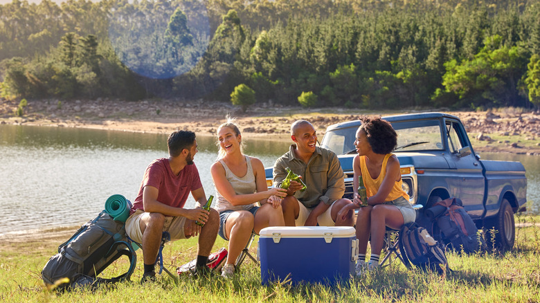 A group of smiling friends drinking beer by a lake with a cooler in front of them