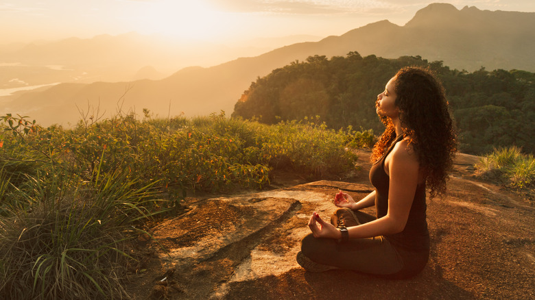 A woman sits in a lotus position on the edge of a forested mountain