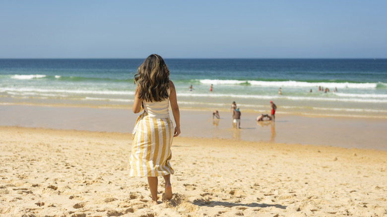 A woman in a dress looking towards the waves while walking on a beach