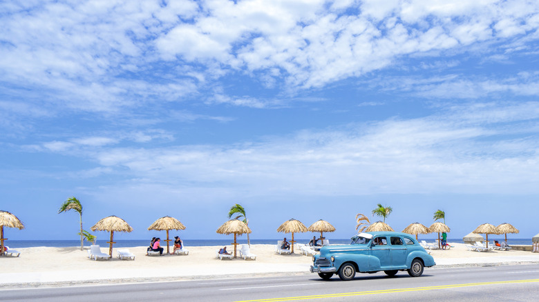 Classic car driving past sandy beach with shaded tables