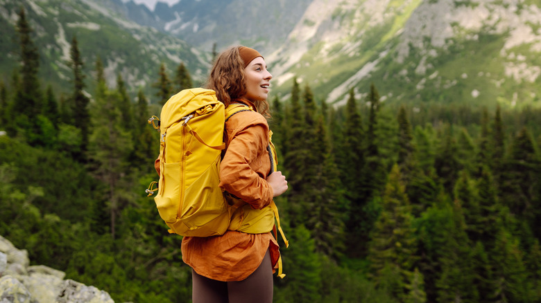 A smiling female traveler with a yellow hiking backpack in front of a mountain vista.