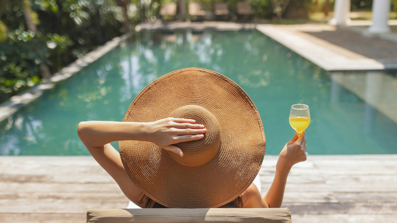 Woman from behind wearing large hat sitting by pool with drink