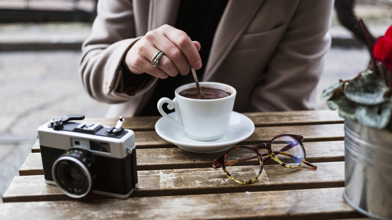A patron stirs coffee at a cafè table as a camera sits nearby