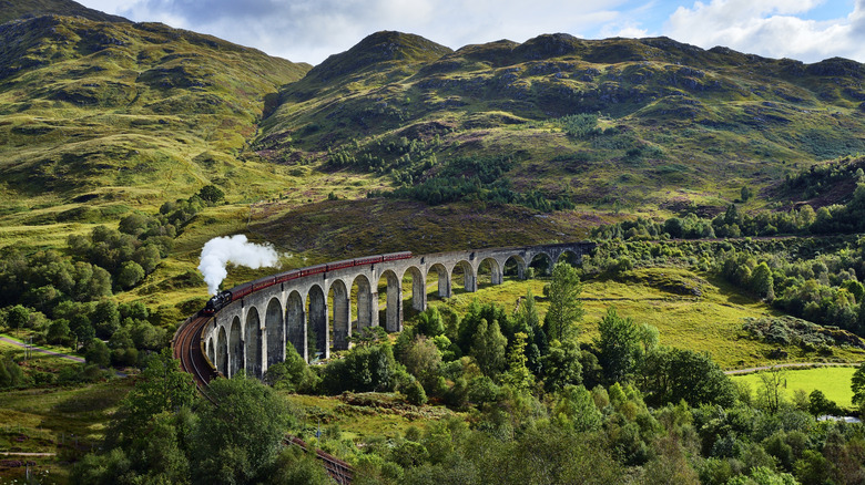 A steam train passes over an arched railway among green hills in Scotland.