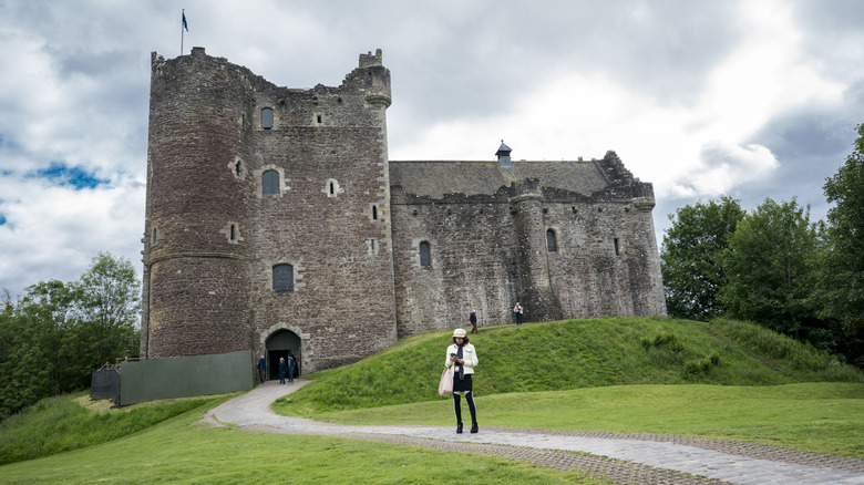 A woman in front of an old castle surrounded by greenery and clouds.