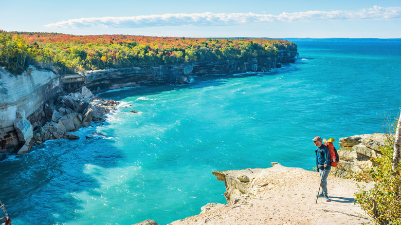 A hiker standing on a cliff with turquoise water and autumn leaves