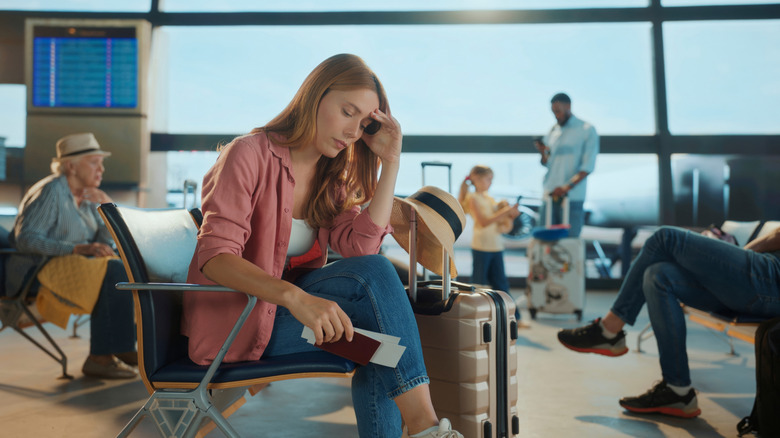 a woman looking fed up sits waiting at an airport with a passport and suitcase