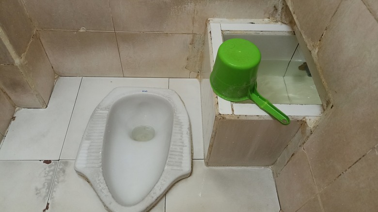 squat toilet with cream colored ceramic walls. Next to the toilet is a water tank with a green dipper.