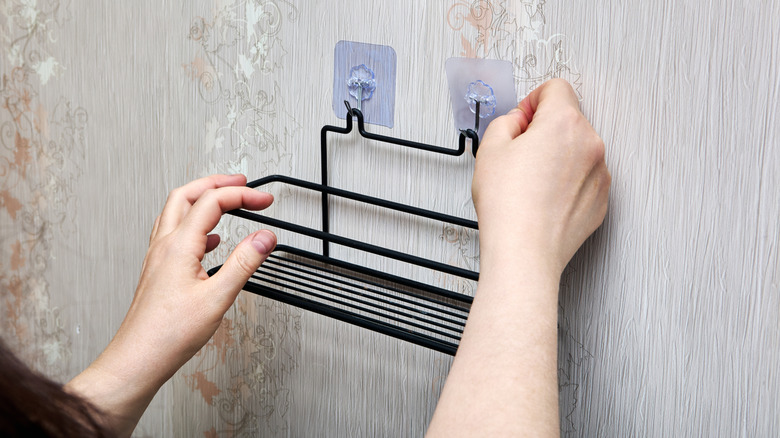 Hands attaching a wire basket against a wall with adhesive hooks.