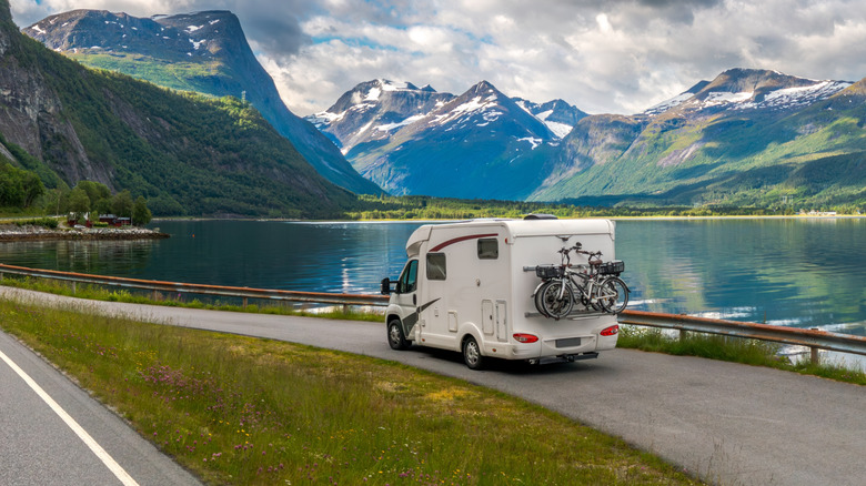 An RV driving on a road with mountains and a lake in the background.