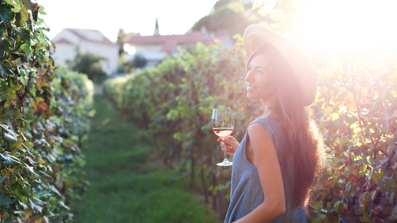 A woman walks through a vineyard with a glass of wine
