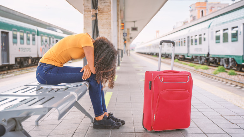 A woman hunched over on a bench with her head on her lap waiting for a delayed train