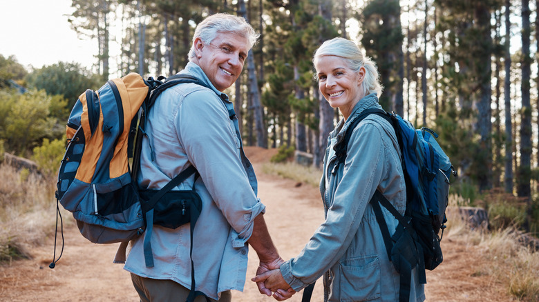 A middle aged couple hiking in a forest holding hands and smiling at the camera.