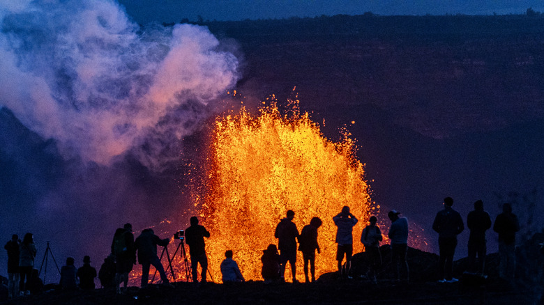 Tourists stand before an active volcano, lava bursting in the air