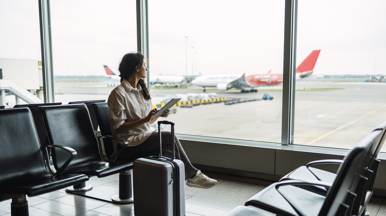 A woman with a tablet and suitcase in an airport waiting area, looking out at the planes.