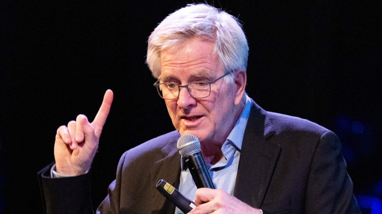 Travel expert Rick Steves with one finger pointed up while the other hand holds a mic