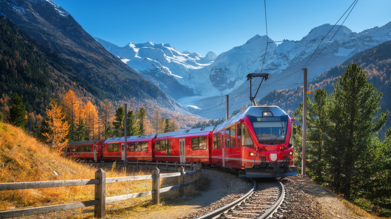 Red modern electric train traveling along a railway, winding through stunning high mountains with snow-capped peaks