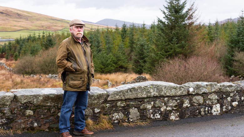 A bearded man wears a waxed jacket near a stone wall
